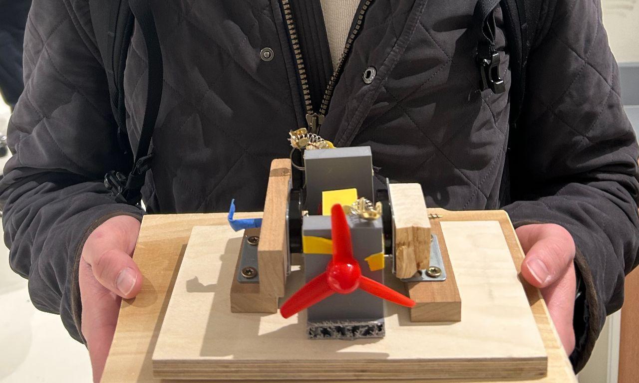 Close view of the completed small wind-driven generator showing its propeller, steel shaft, rotating 80-turn coil, opposing magnets, brush contacts, 3D-printed supports, and wooden base.